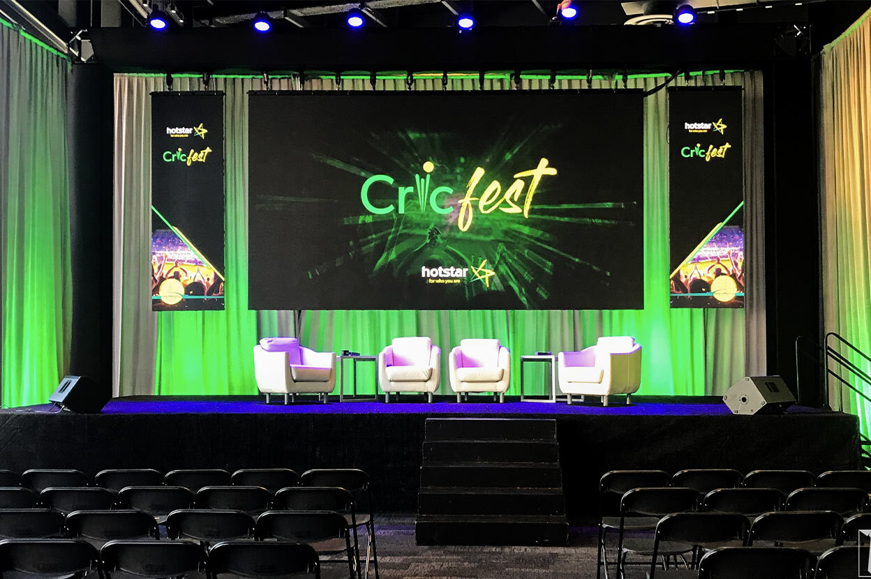 Stage with green accent lighting, Cricfest banners, and white armchairs.