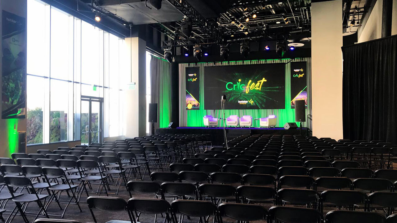 Many rows of chairs in front of a stage containing comfortable chairs and Cricfest banners. The room has green accent lighting.